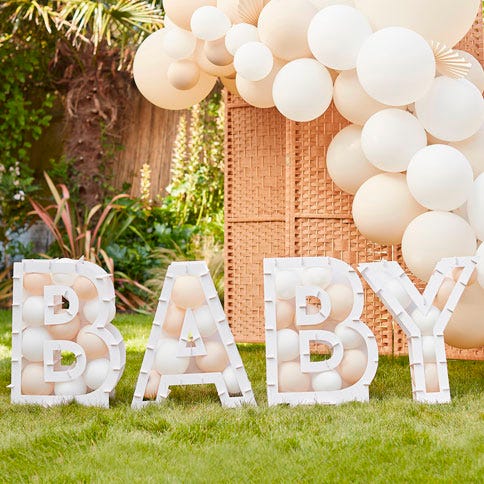 Neutral coloured balloon mosaic letters spelling out baby on the grass outside, surrounded by a balloon arch in the same colours. There is a rattan divider behind the arch and some garden plants to the left.