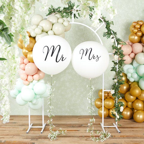 A white metal wedding arch decorated with balloons and foliage, in the centre of the arch is two giant white balloons with 'mr and mrs' written in cursive, to the right of the arch is a gold, green and blush balloon column and to the left is more foliage.