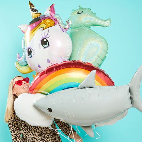 A giant unicorn balloon, a seahorse balloon, a rainbow balloon and a shark balloon held in a bunch by a blonde woman wearing pink heart sunglasses, who is looking up at the balloons. The image is on a turquoise background.