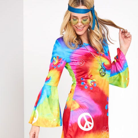 A blonde woman on a white background wearing a 60s-style tie dye swing dress with peace symbols on it, she's wearing a long chain peace symbol necklace, green circle sunglasses, a blue head tie looking down to the floor